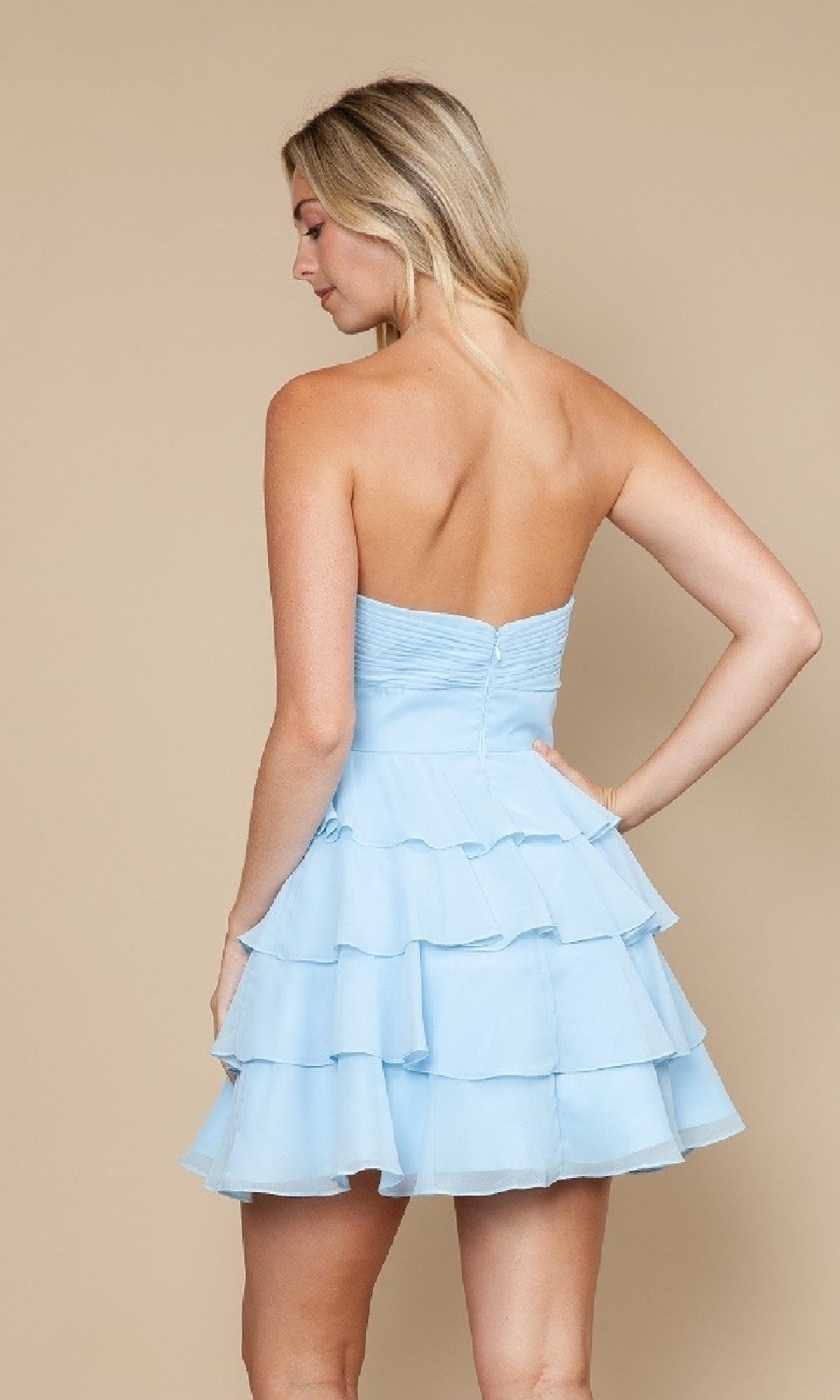 A woman in the Poly USA 9802 Cut-Out Strapless Short Hoco Dress, light blue with tiered ruffles, stands with her back to the camera against a beige background.